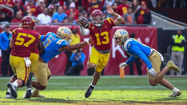 USC quarterback Caleb Wiliams, wearing a maroon jersey with yellow pants and holding the football aloft, jumps through two UCLA players, decked out in baby blue and gold, in front of a large crowd.